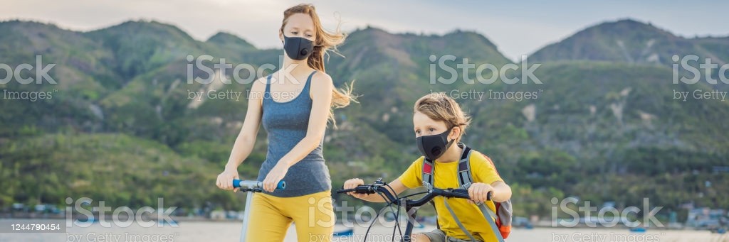 Active school kid boy and his mom in medical mask riding a bike with backpack on sunny day. Happy child biking on way to school. You need to go to school in a mask because of the coronavirus epidemic. BANNER, LONG FORMAT